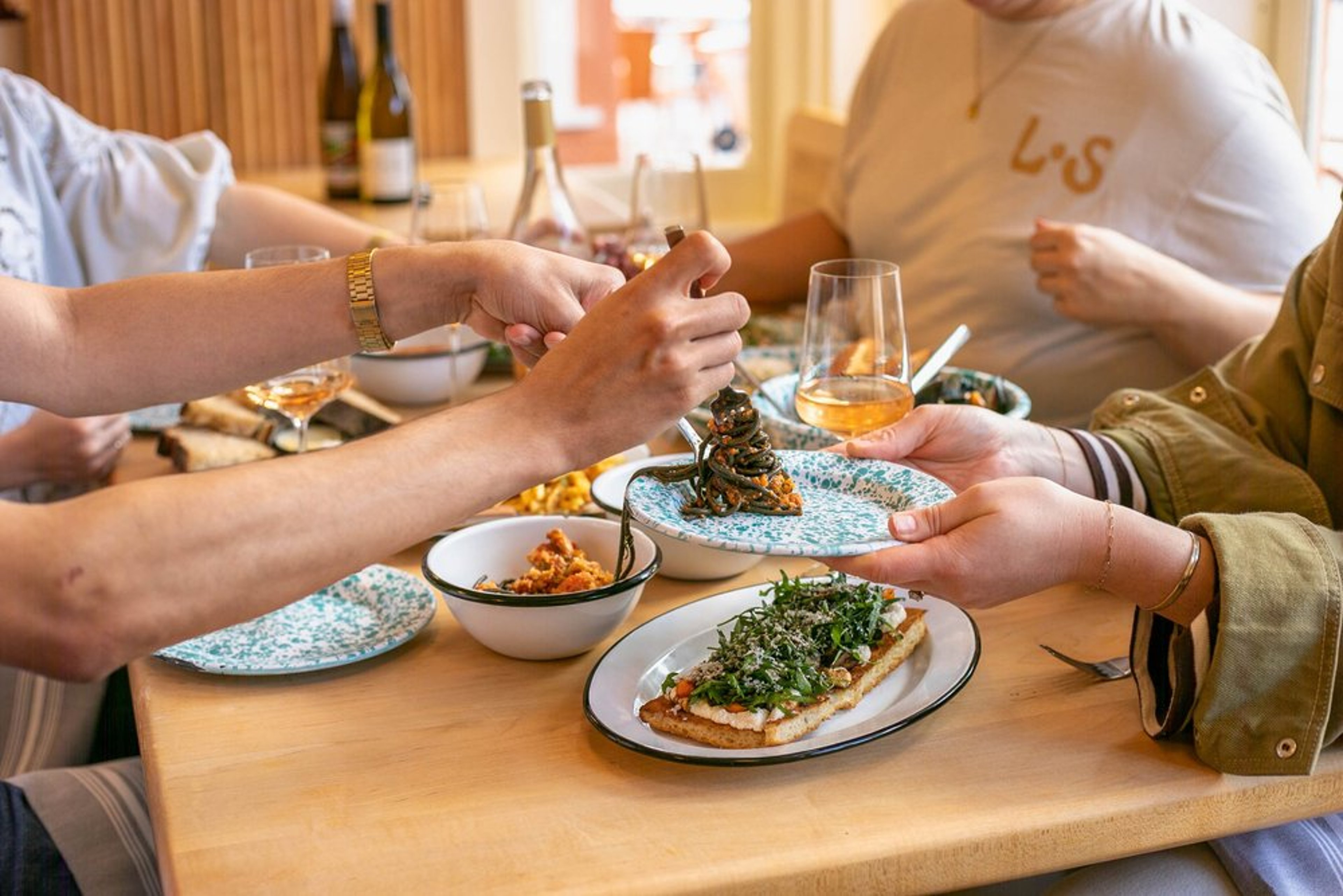 People sharing a meal at a dining table with dishes including pasta and a glass of rosé wine.